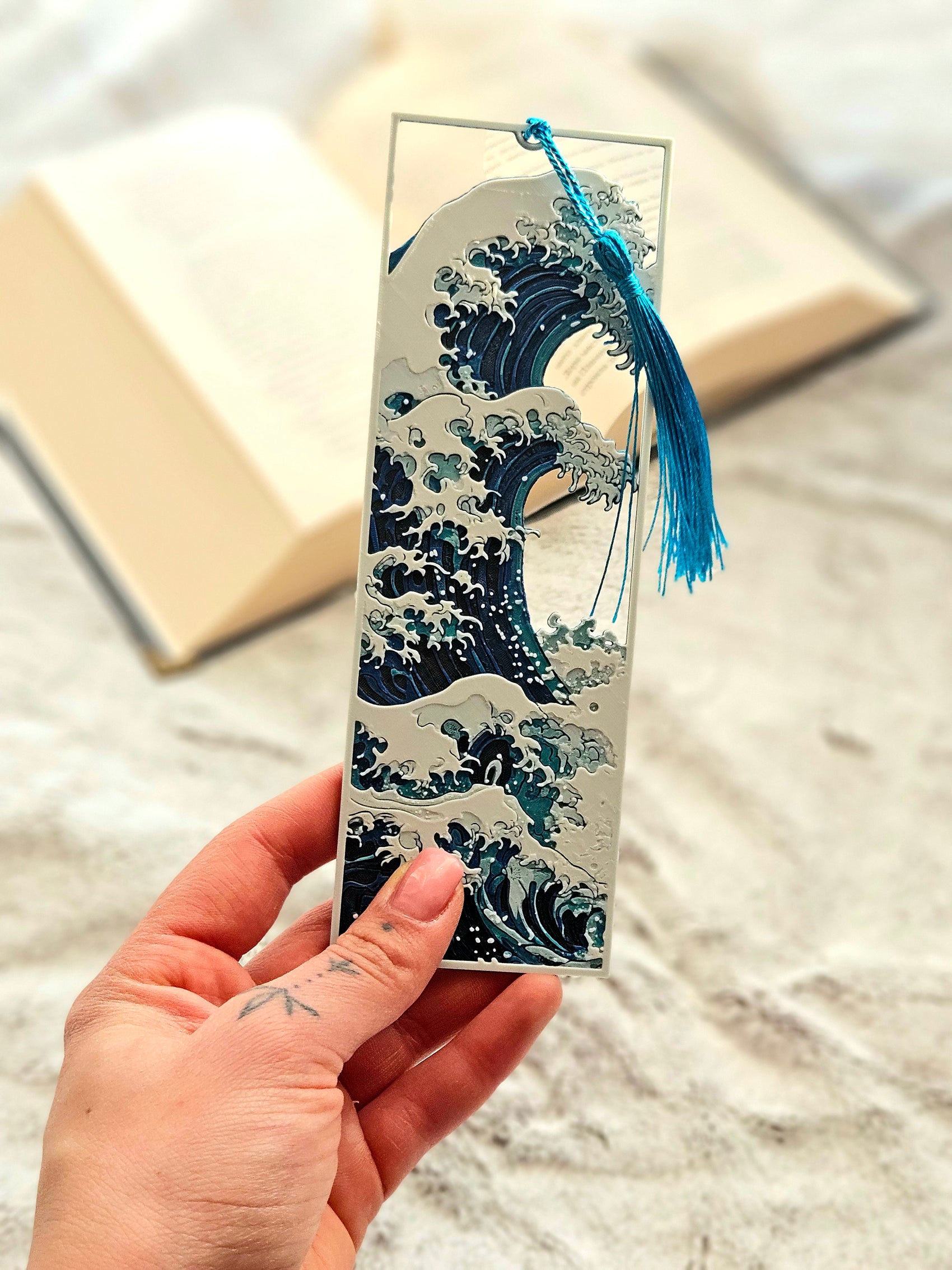 Hand holding a decorative bookmark with wave design and blue tassel.